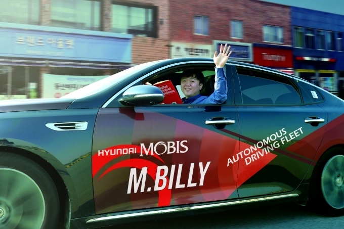 서산주행시험장내 도로에서 자율주행차 M.BILLY를 테스트하는 연구원이 책을 보며 밖으로 손을 흔들고 있다.
